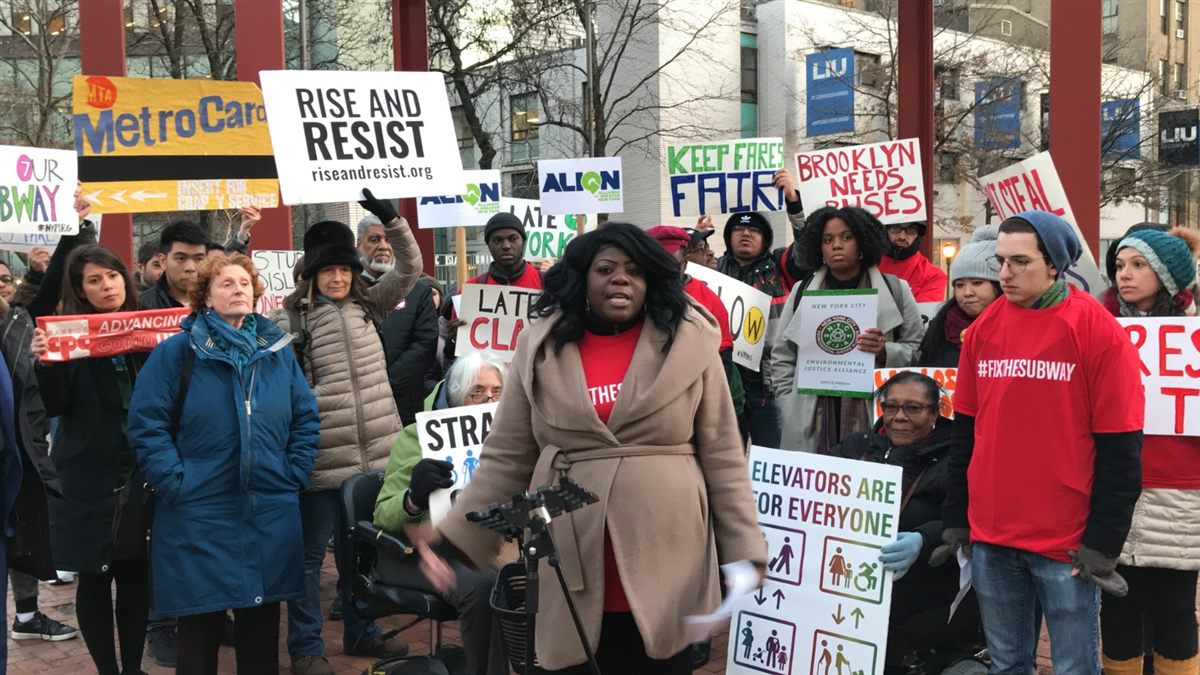 Danna Dennis speaking at a Fair Fares NYC rally, with a "Keep Fares Fair" sign visible behind her.
