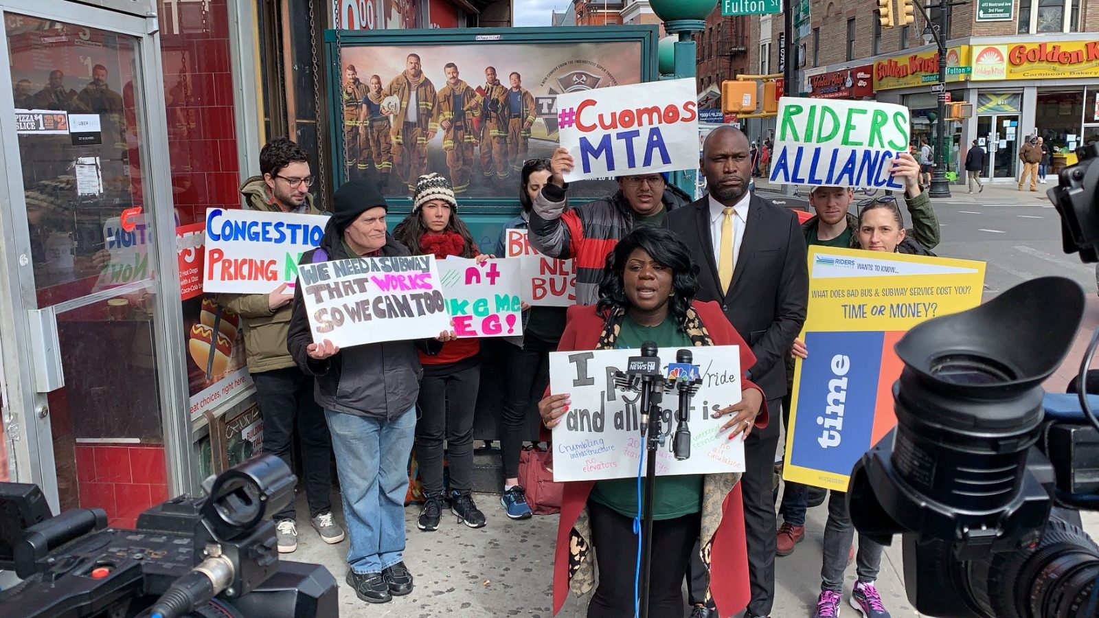 Danna Dennis speaking at a press conference surrounded by advocates and news cameras, part of a transit advocacy campaign.